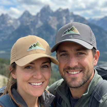 Mann und Frau tragen beige und graue Outdoor-Kappen mit Berg-Stickerei beim Wandern vor einer Gebirgskulisse.