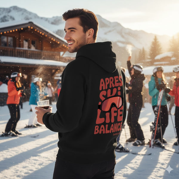 Lächelnder Mann im Schnee hält Glühwein und trägt einen schwarzen bawearia Hoodie mit dem roten Rückenprint "APRES SKI LIFE BALANCE" und Skibrille-Motiv.