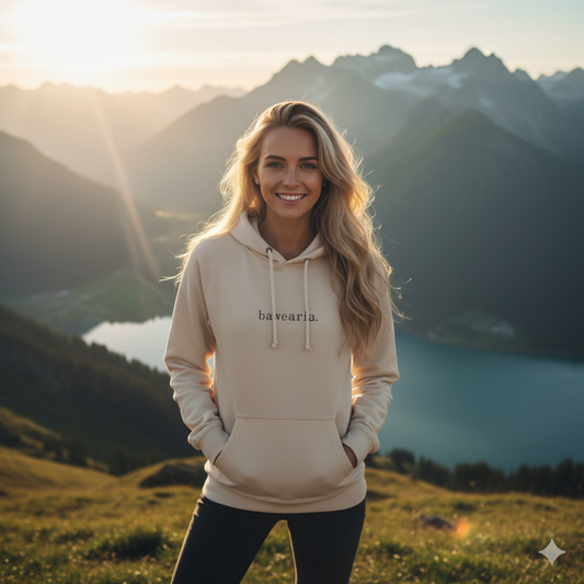 Frau steht lächelnd vor Bergpanorama mit See und trägt einen beigen bawearia Hoodie mit Logo-Schriftzug auf der Brust