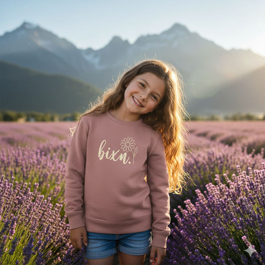 Lachendes Mädchen steht in einem lila Blumenfeld vor Bergkulisse und trägt ein altrosa Sweatshirt mit dem Schriftzug "bixn." und einer Blumen-Grafik