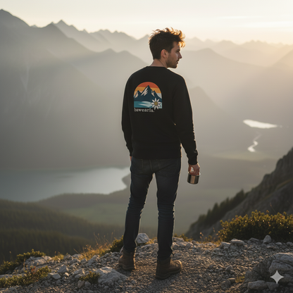 Mann steht bei Sonnenuntergang auf einem Berggipfel, hält einen Becher in der Hand und trägt ein schwarzes Sweatshirt mit buntem Retro-Berg-Rückenprint und Edelweiß
