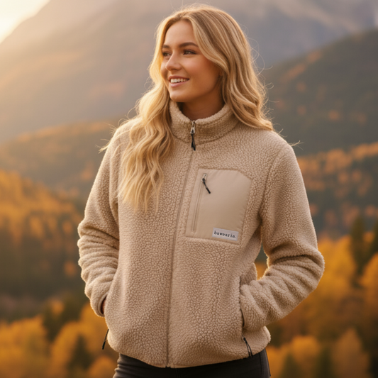 Frau steht vor herbstlichem Bergpanorama und trägt eine beige Teddy-Fleece-Jacke mit Brusttasche und bawearia-Logo
