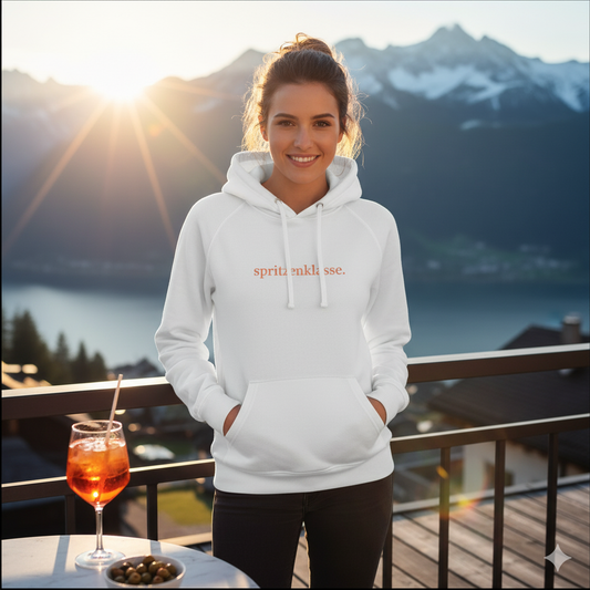 Vorderseite eines weißen Damen-Hoodies mit dem orangen Schriftzug "spritzenklasse." getragen vor einem sonnigen Bergpanorama mit Aperol Spritz im Vordergrund.