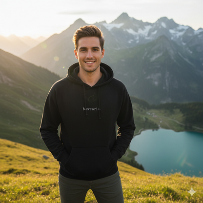 Herr steht lächelnd vor Bergpanorama mit See und trägt einen beigen bawearia Hoodie mit Logo-Schriftzug auf der Brust